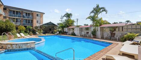 A swimming pool with lounge chairs and a covered seating area, surrounded by buildings and palm trees.