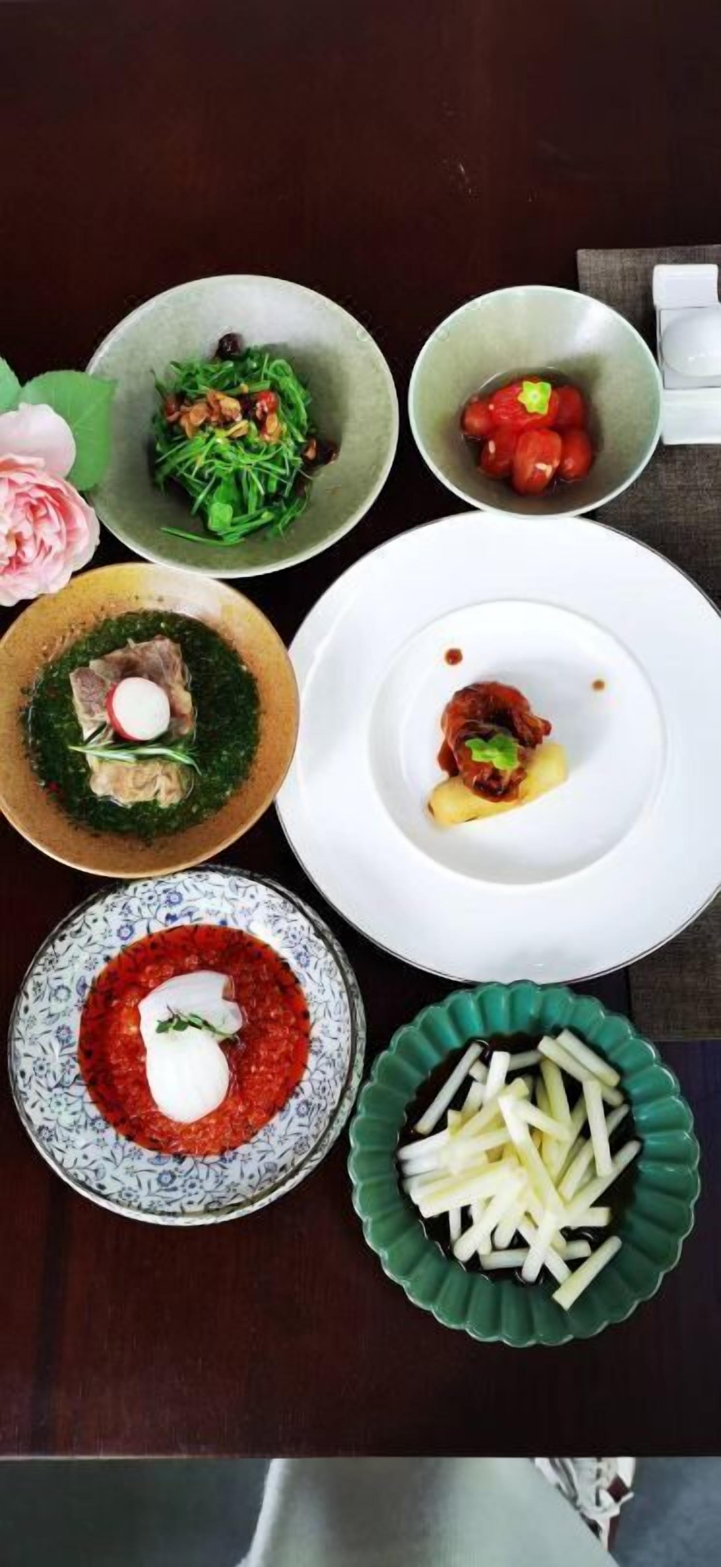 A table with various dishes including a salad, a bowl of tomatoes, a dish with meat and greens, a plate with a red sauce, and a bowl of sliced potatoes.