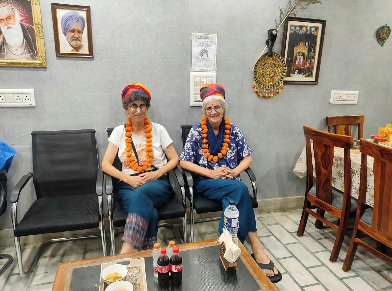 Two individuals seated on black chairs in a room with a tiled floor, a wooden table with bottles, and framed pictures on the wall.