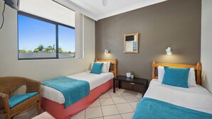 A room with two beds, a small table, a chair, a ceiling fan, and a window with a view of palm trees.