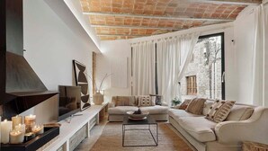 A living room with a sofa, a coffee table, and a TV on a stand. There are candles on a shelf and a window with curtains.