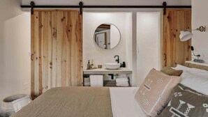 A bedroom with a wooden wardrobe, a round mirror, a bedside table with a lamp, and a bed with patterned pillows.