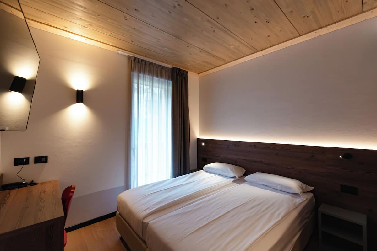 Habitación de hotel con techo de madera, cama con ropa blanca, escritorio con silla y televisor montado en la pared.