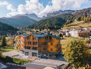 Un edificio moderno de madera con balcones, ubicado en un pueblo montañoso con casas y una iglesia al fondo.