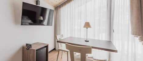 Una habitación compacta con techo de madera, un televisor de pantalla plana montado en la pared, un mueble de madera pequeño, una mesa con una silla y cortinas traslúcidas en la ventana.