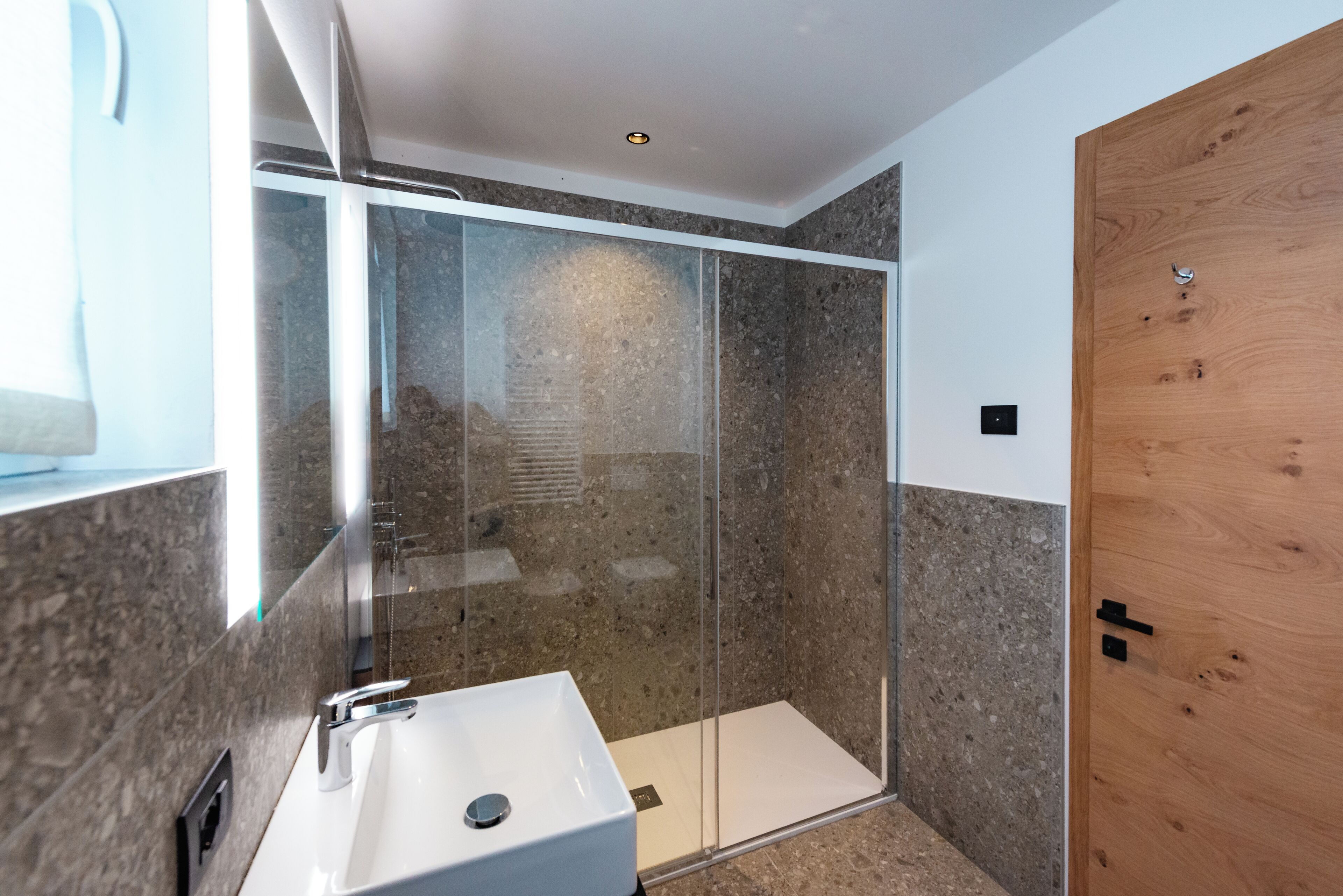 A modern bathroom with a glass-enclosed shower, a white sink, and a wooden door.
