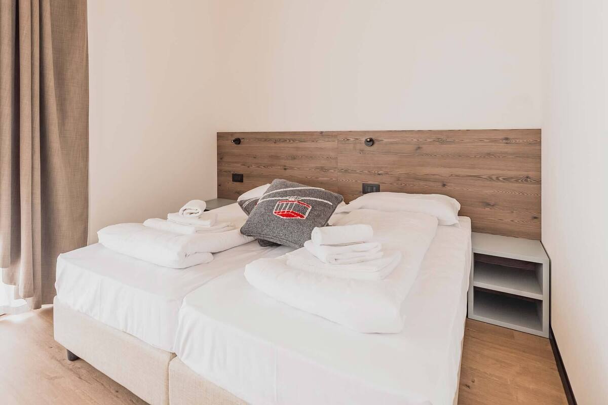 Una cama bien tendida con sábanas blancas y un almohadón gris con un logotipo rojo, un cabecero de madera y una mesita de noche con un cajón blanco.