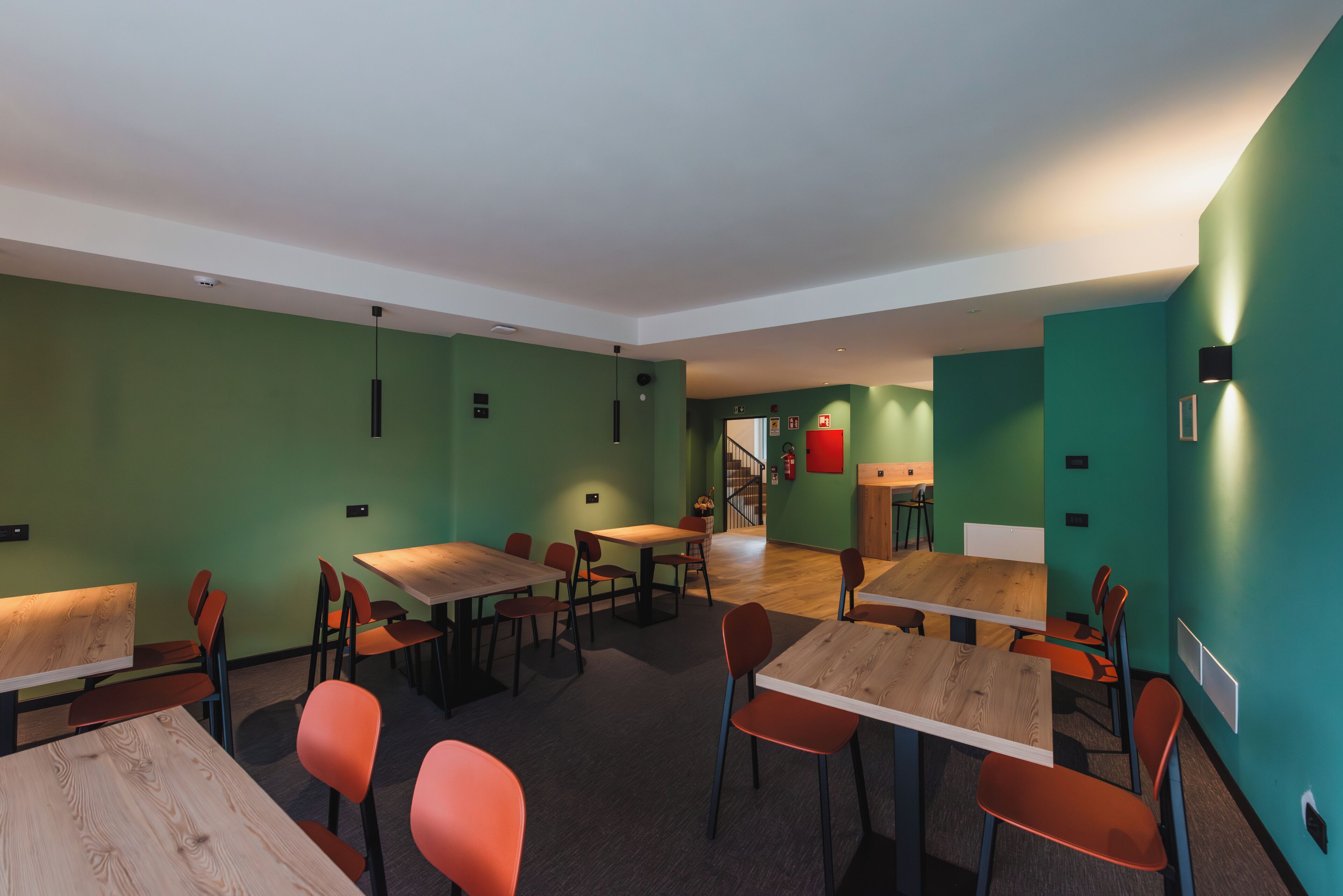 A modern dining area with wooden tables and orange chairs, green walls, and a staircase in the background.