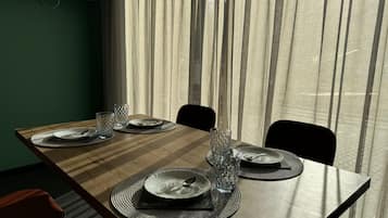 A dining table set with plates, glasses, and cutlery, with a view of sheer curtains and a green wall in the background.