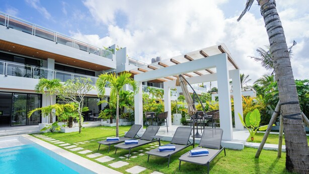 A modern outdoor pool area with lounge chairs and a pergola, surrounded by greenery and a multi-level building. - Gandum Uluwatu Luxury Boutique Stay - Gandum Villa Complex (Pecatu)