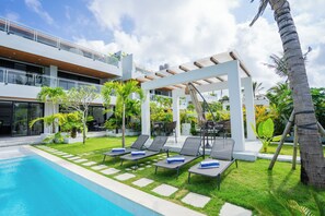 Uma área moderna de piscina ao ar livre, com espreguiçadeiras e um pergolado, cercada por vegetação e um edifício em vários níveis.