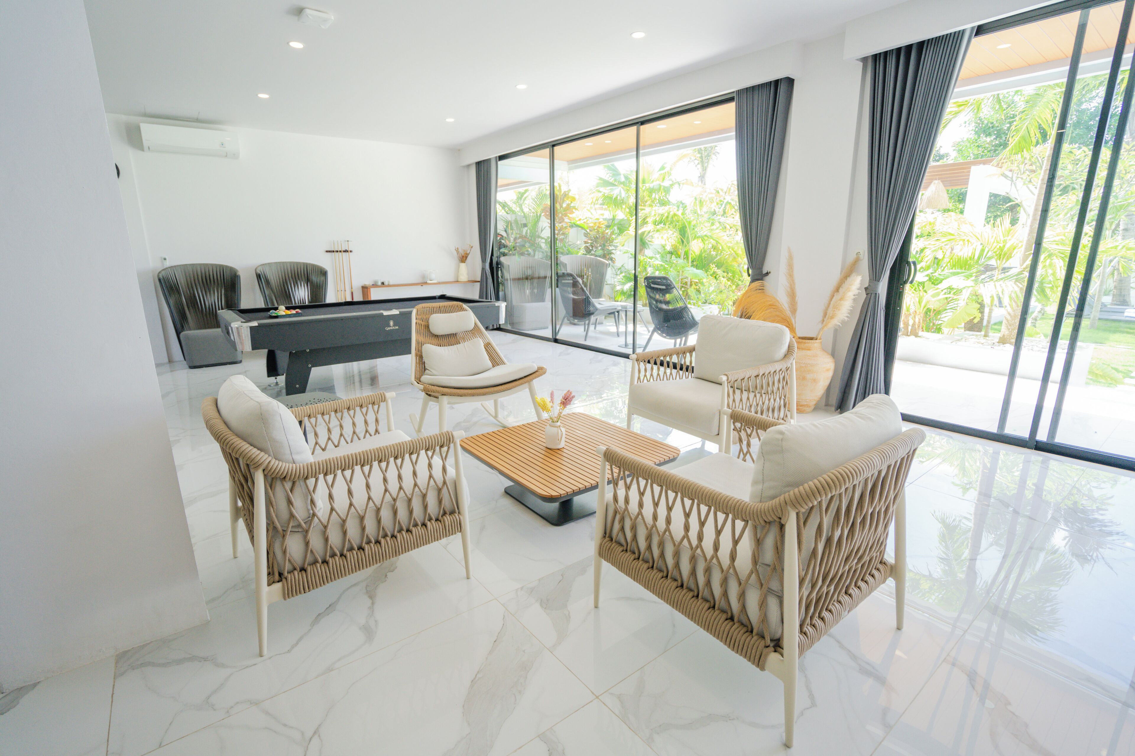 A modern living room with wicker furniture, a pool table, and large windows offering a view of the outdoors.
