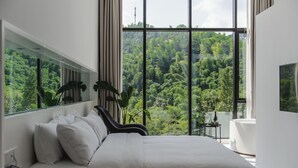 Un dormitorio moderno con una cama grande, una mesita de noche con un teléfono y vistas a un bosque frondoso a través de amplias ventanas.
