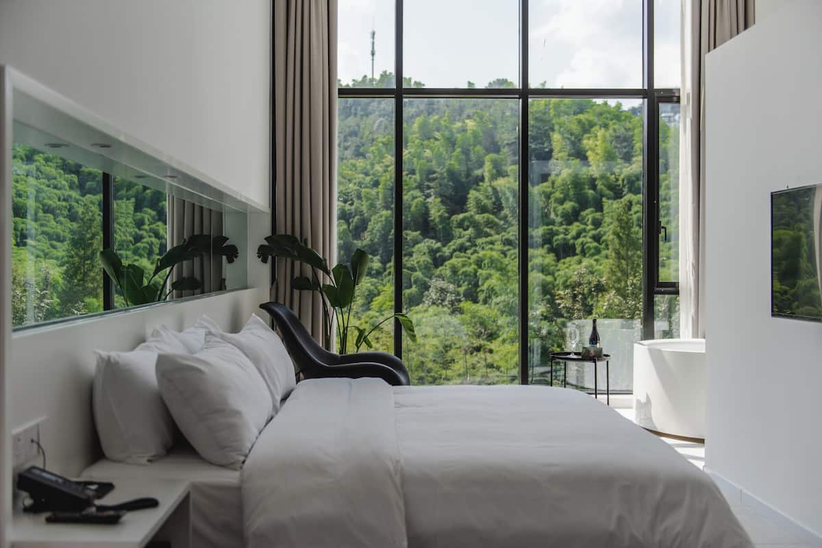 Une chambre moderne avec un grand lit, une table de chevet avec un téléphone et une vue sur une forêt luxuriante, visible à travers de grandes fenêtres.