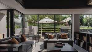 Una sala de estar moderna con un sofá, una mesa de centro y una estantería. Se ve una ventana grande con vistas a un jardín y mobiliario de exterior.