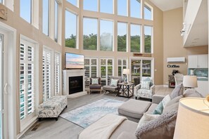 A spacious living room with a fireplace, comfortable seating, large windows, and a view of the outdoors.