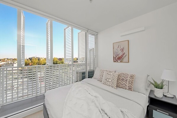 A bedroom with a large window, a bed with patterned pillows, a bedside table with a lamp, and a framed artwork on the wall.