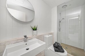 Une salle de bain moderne avec un lavabo blanc, des toilettes, un espace douche et un miroir rond.