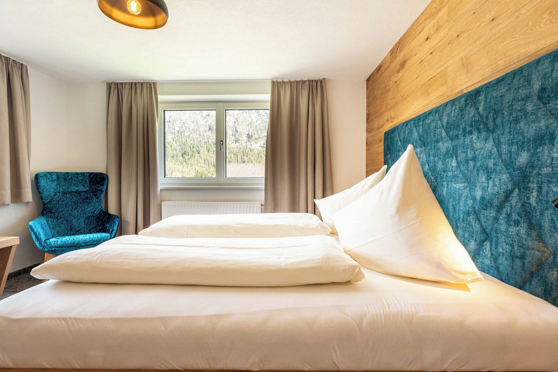 Una habitación de hotel con cama, una silla azul, una ventana con cortinas y un cabecero de madera.
