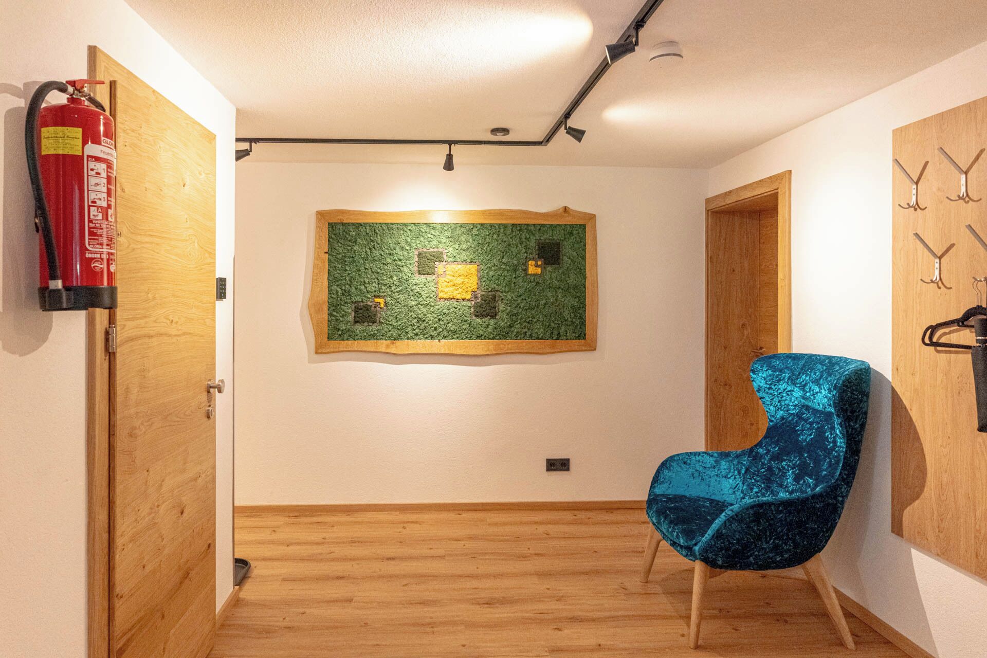 Una habitació amb sòl de fusta, una cadira blava, una obra d'art mural verda i un extintor fixat a la paret.
