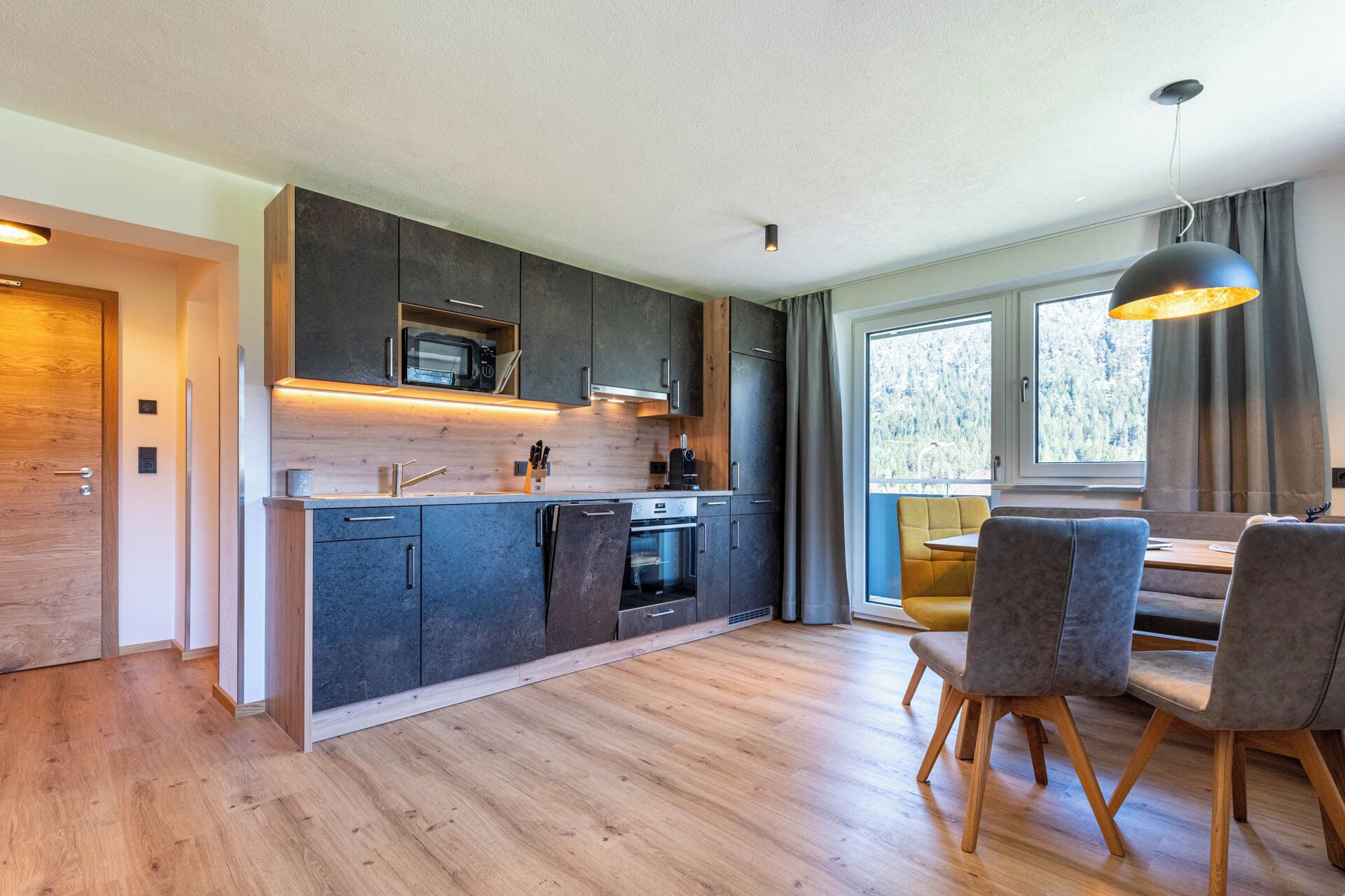 A modern kitchen with dark cabinets, a wooden countertop, and a built-in microwave. There is a dining area with a table and chairs, and a large window providing natural light.