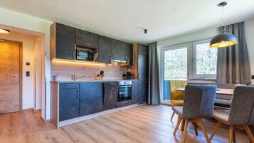 A modern kitchen with dark cabinets, a wooden countertop, and a built-in microwave. There is a dining area with a table and chairs, and a large window providing natural light.