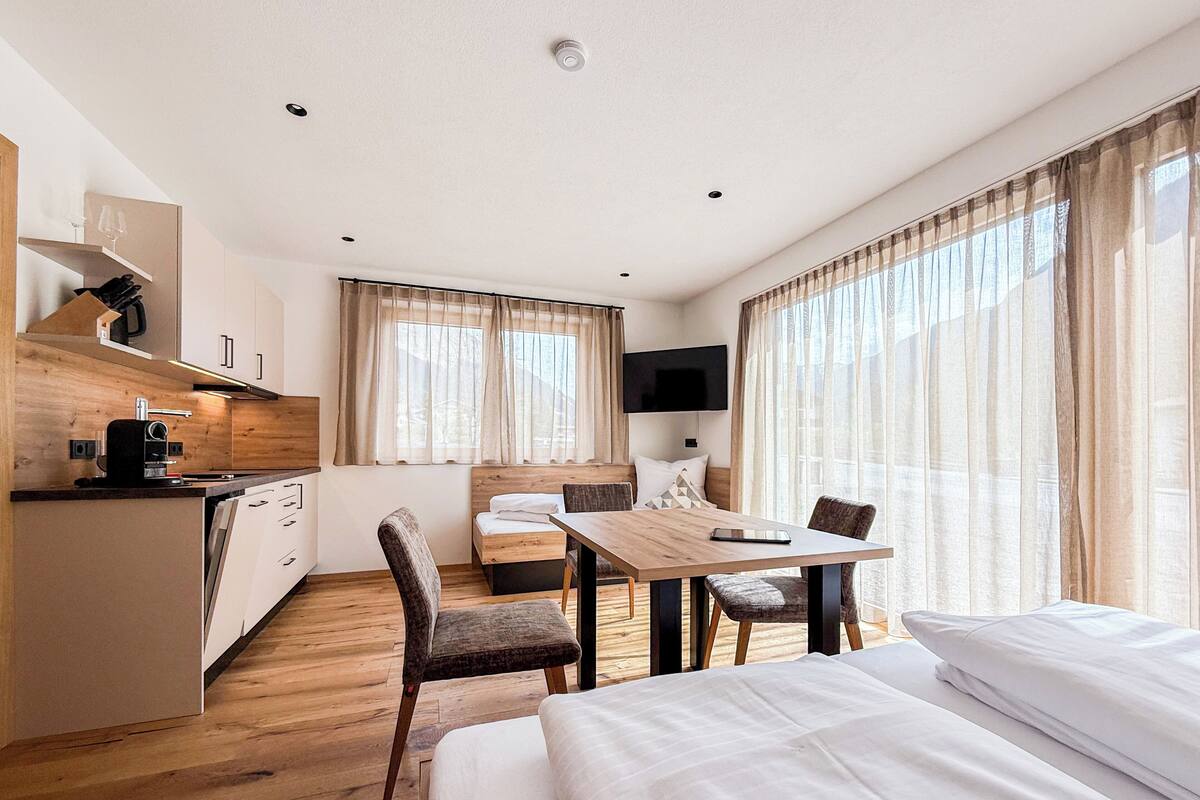 A modern hotel room with a wooden floor, a dining table, a bed, a TV, and a kitchen area with a coffee machine.