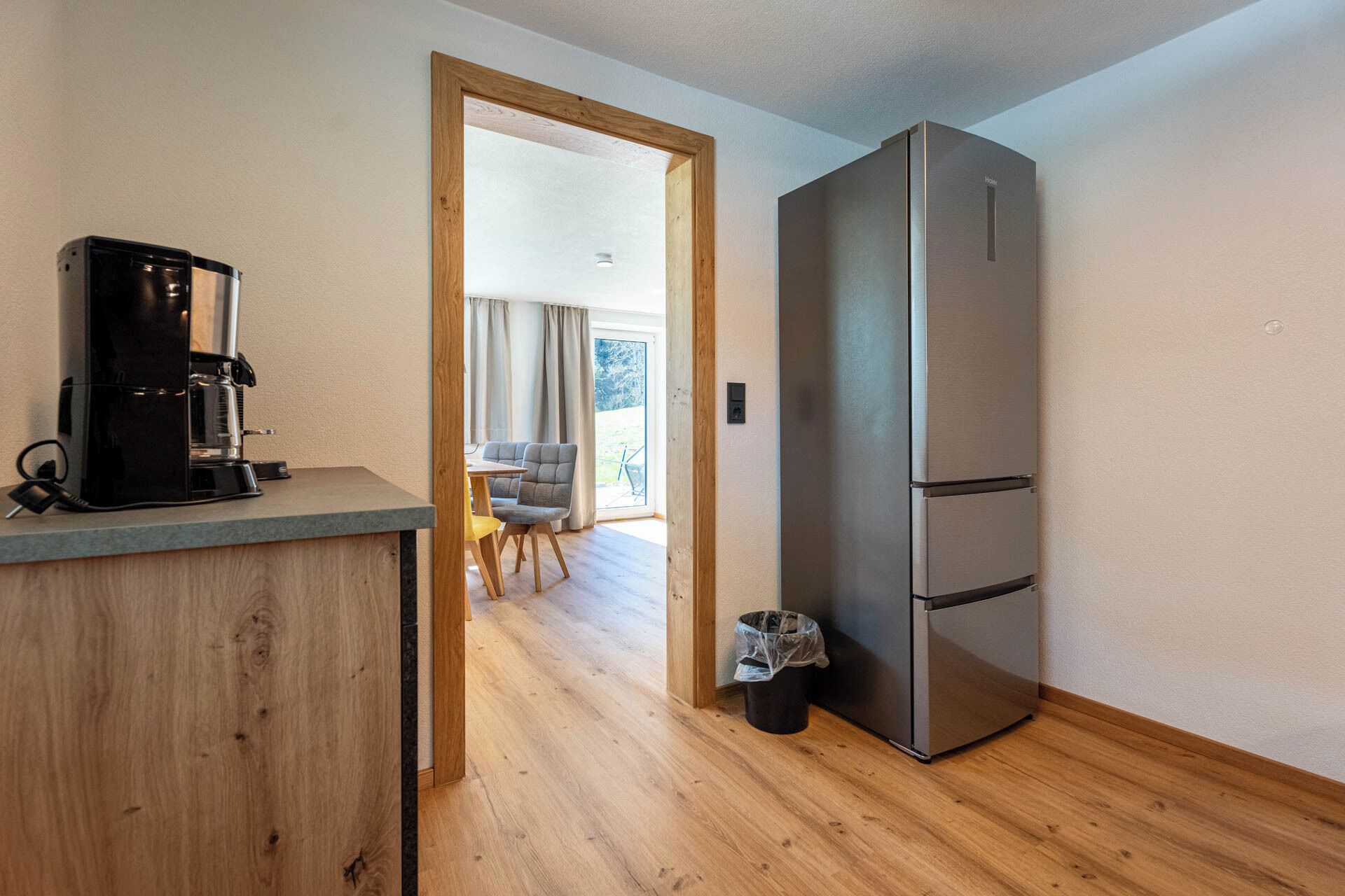 A compact kitchen with a coffee machine, a stainless steel refrigerator, and a wooden floor.