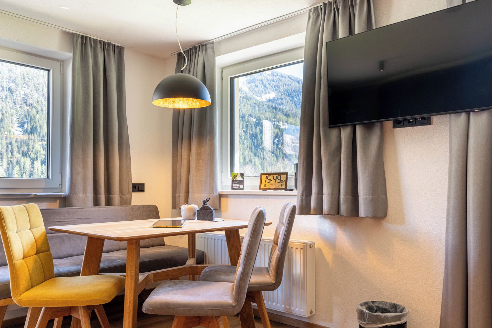 A cozy dining area with a wooden table and cushioned chairs, a flat-screen TV mounted on the wall, and a window with a view of a snowy mountain.