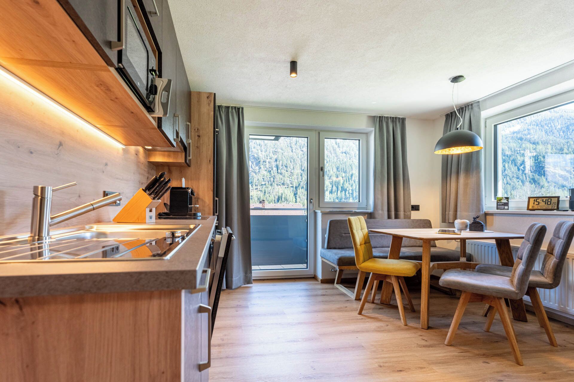 Een moderne keuken met een houten aanrecht en roestvrijstalen apparatuur, een eethoek met tafel en stoelen, en grote ramen met uitzicht op de omgeving.