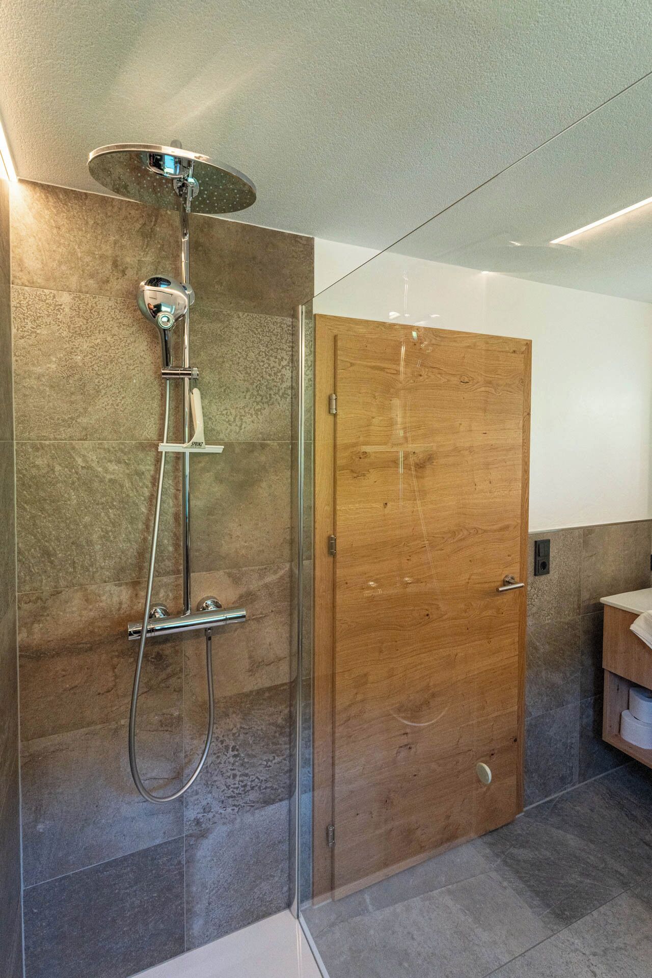 Un baño moderno con ducha envuelta en cristal, puerta de madera y piso de baldosas.