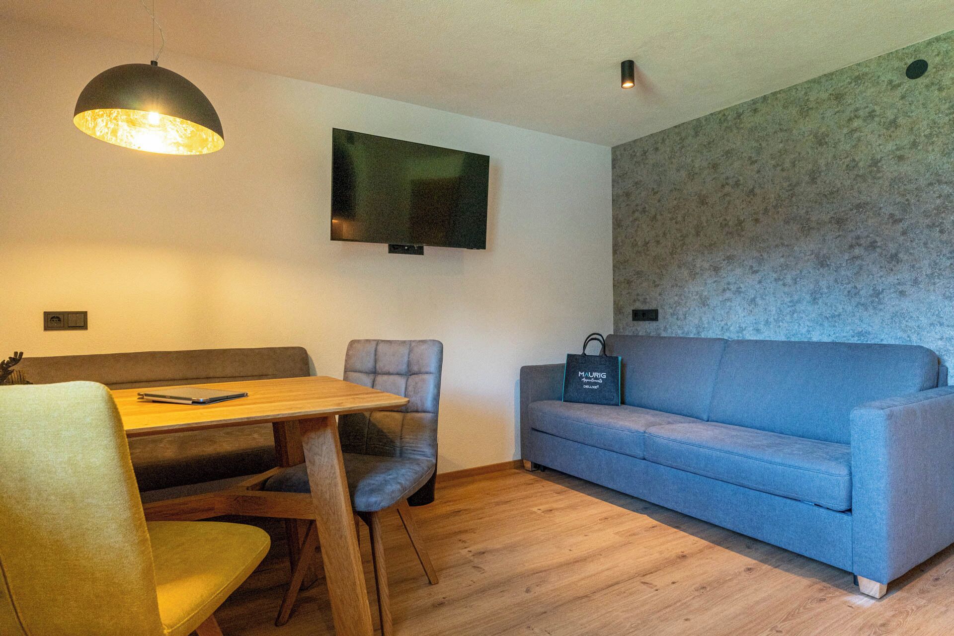 A small, modern living space with a wooden dining table, two chairs, a blue sofa, and a mounted flat-screen TV.