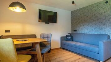 A small, modern living space with a wooden dining table, two chairs, a blue sofa, and a mounted flat-screen TV.