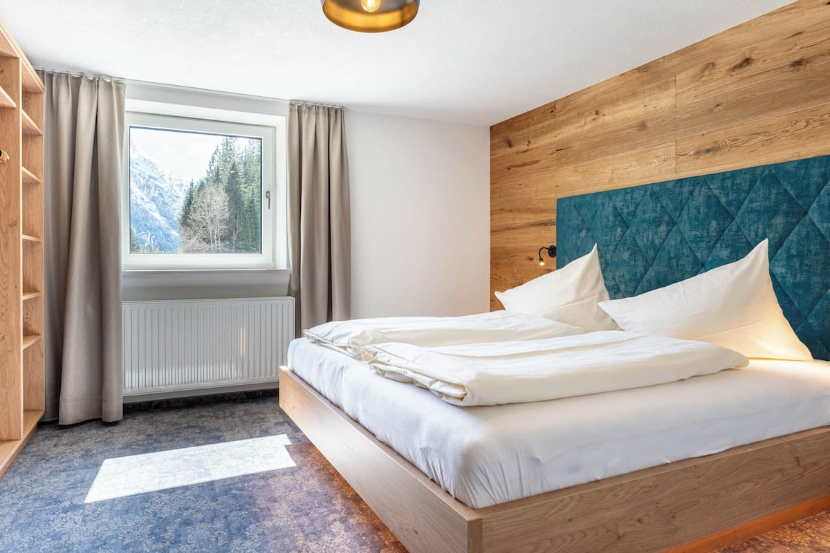 A modern bedroom with a wooden headboard, a large window with curtains, and a radiator.