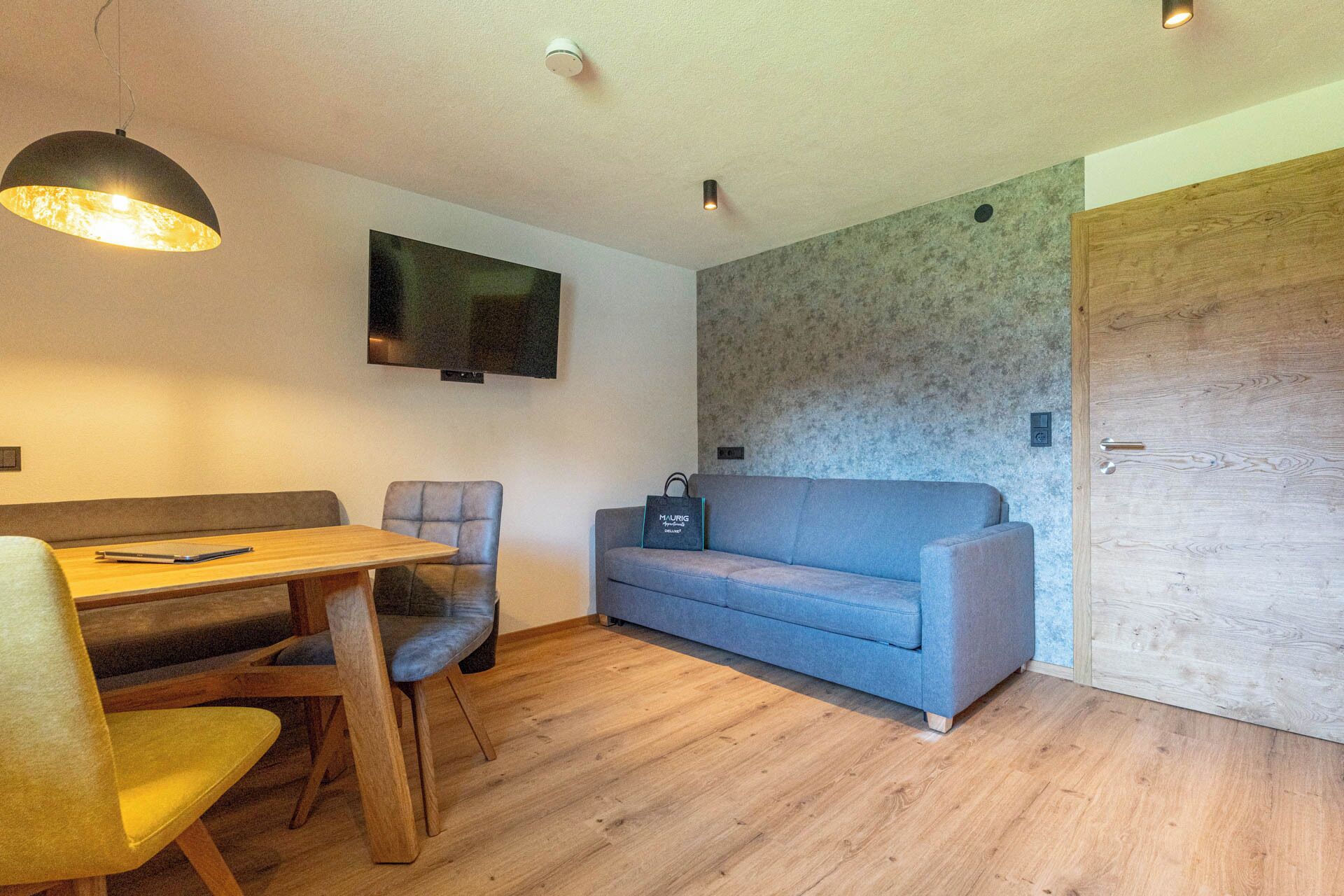 A compact living space with a wooden dining table, two chairs, a sofa, and a television mounted on the wall.