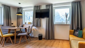 A modern living room with a wooden floor, a sofa, a dining table with chairs, a TV mounted on the wall, and a window with curtains. - Maurig Appartement (Elbigenalp)