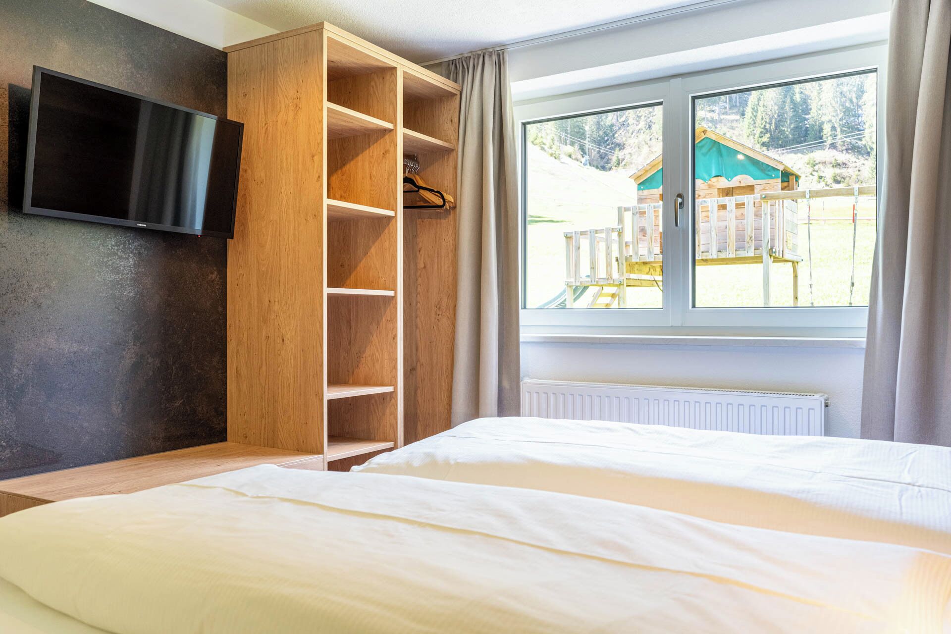 Una camera da letto con un letto, un armadio in legno, una televisione a parete e una finestra vista su una struttura in legno.