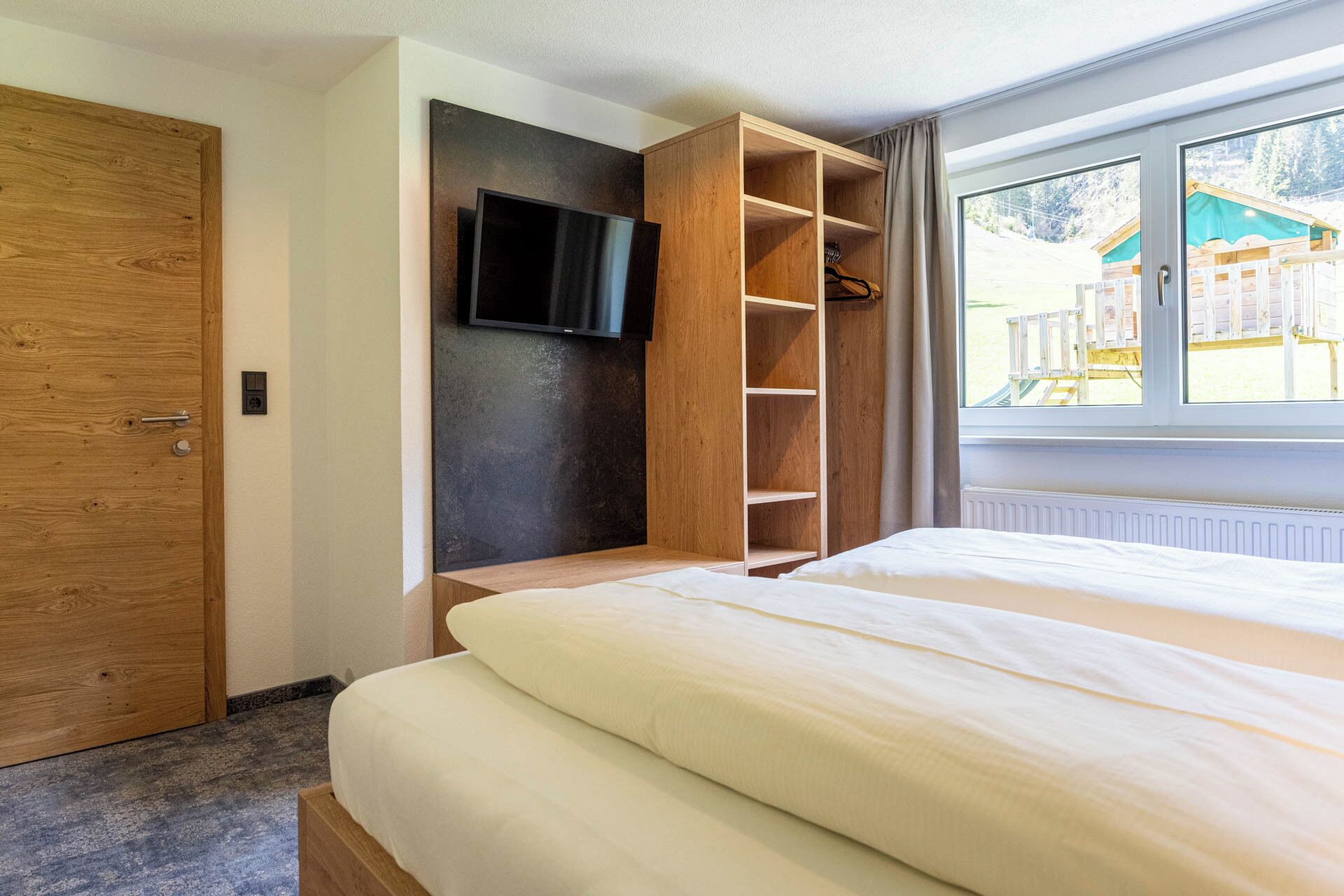 Una camera da letto con un letto, un armadio in legno, una TV a parete e una finestra che dà su un edificio.