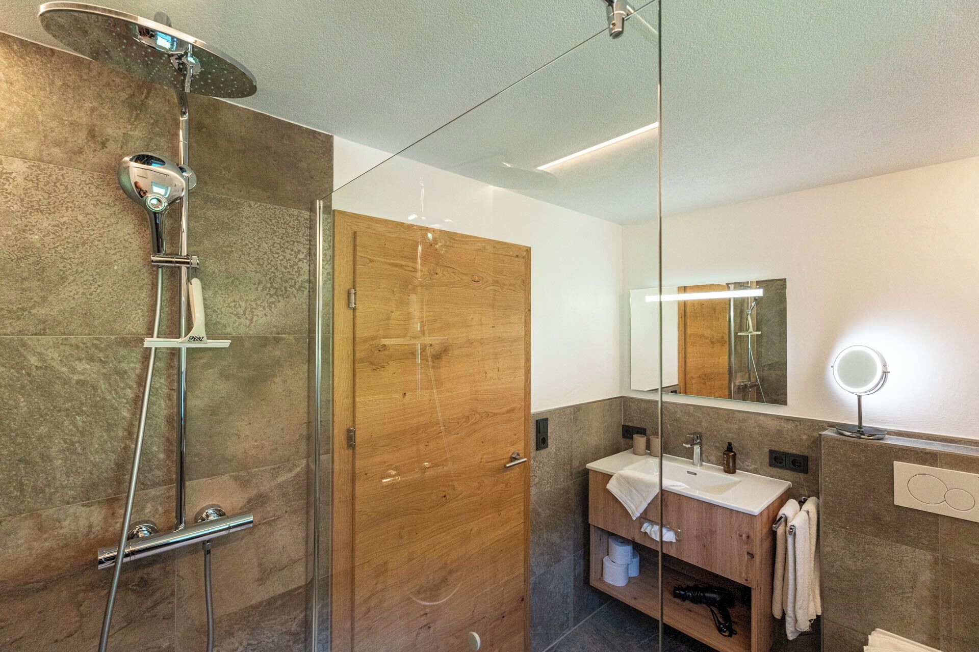 Un baño moderno con ducha envuelta en vidrio, puerta de madera, tocador con espejo y perchero para toallas.