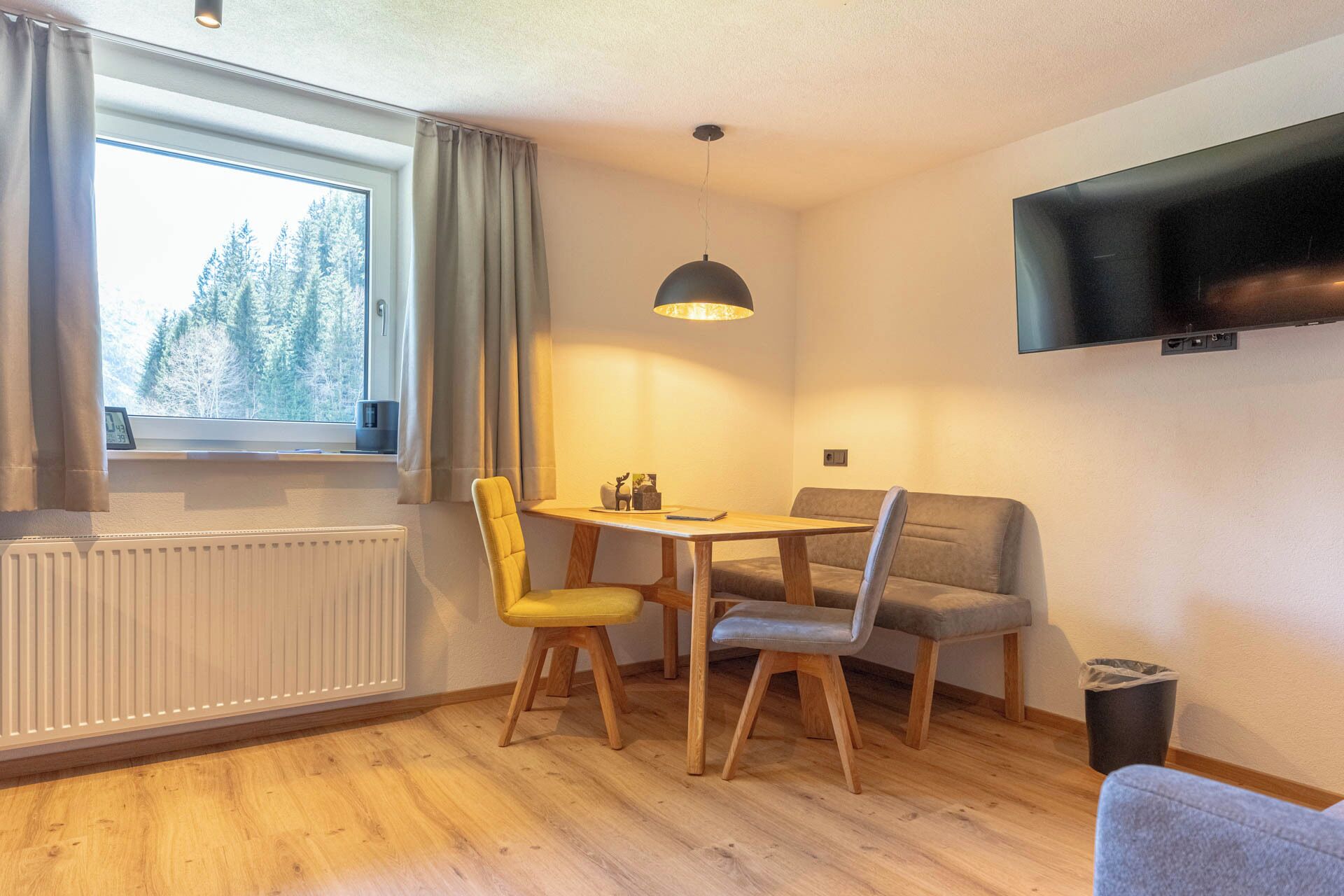 Um quarto acolhedor com piso de madeira, uma mesa de jantar com cadeiras, um sofá, uma televisão fixada na parede e uma janela com cortinas.
