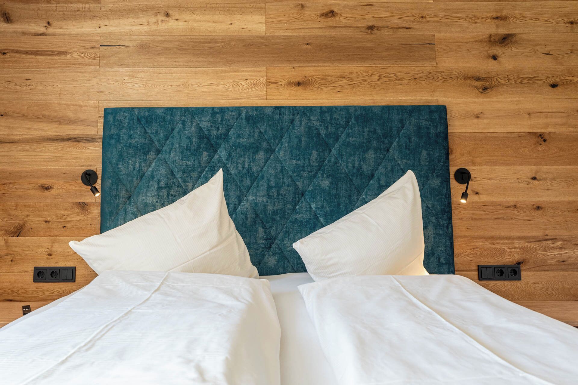 A bed with white linens and a teal headboard against a wooden wall.