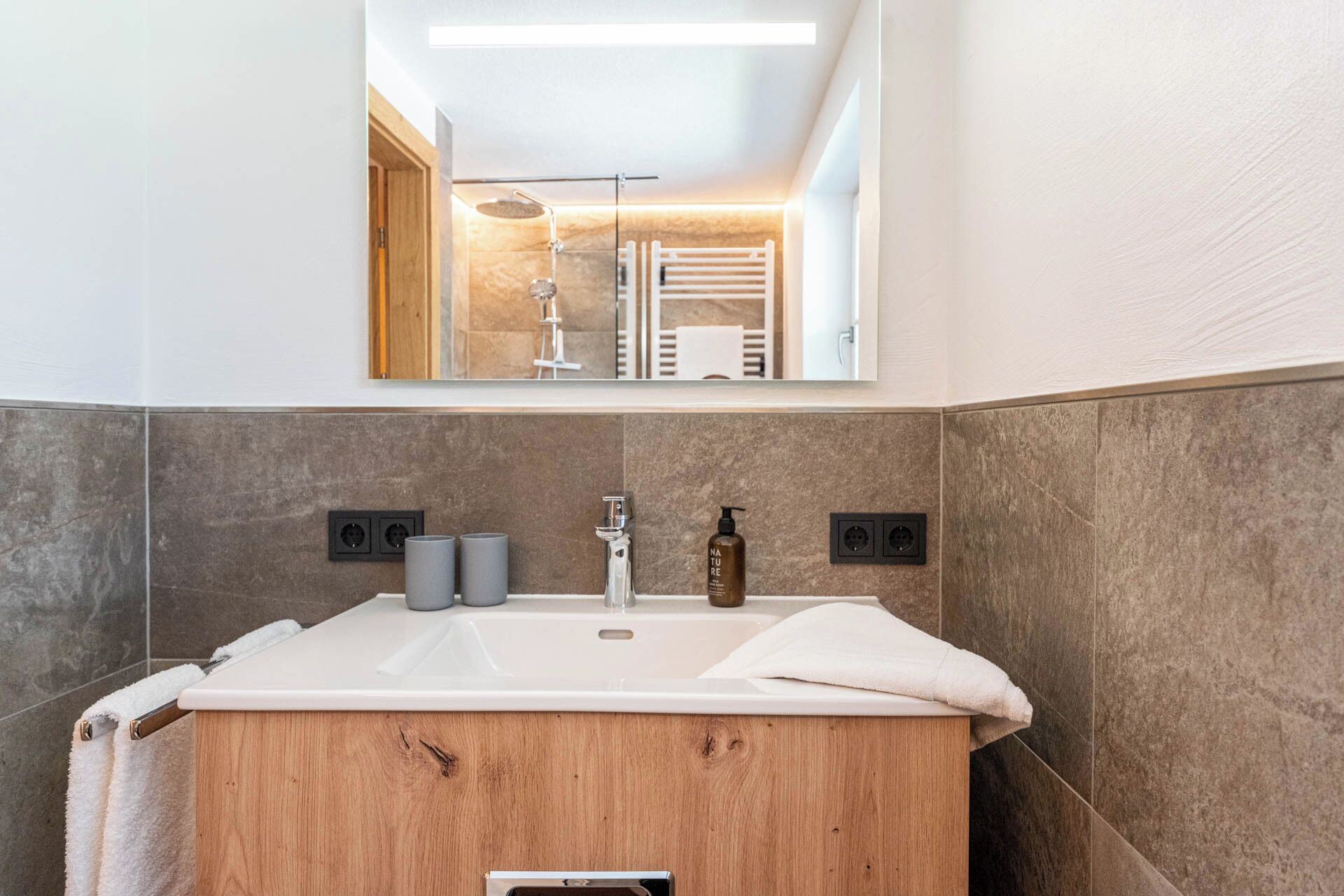 A modern bathroom with a wooden vanity, a large mirror, and a shower area visible in the reflection.