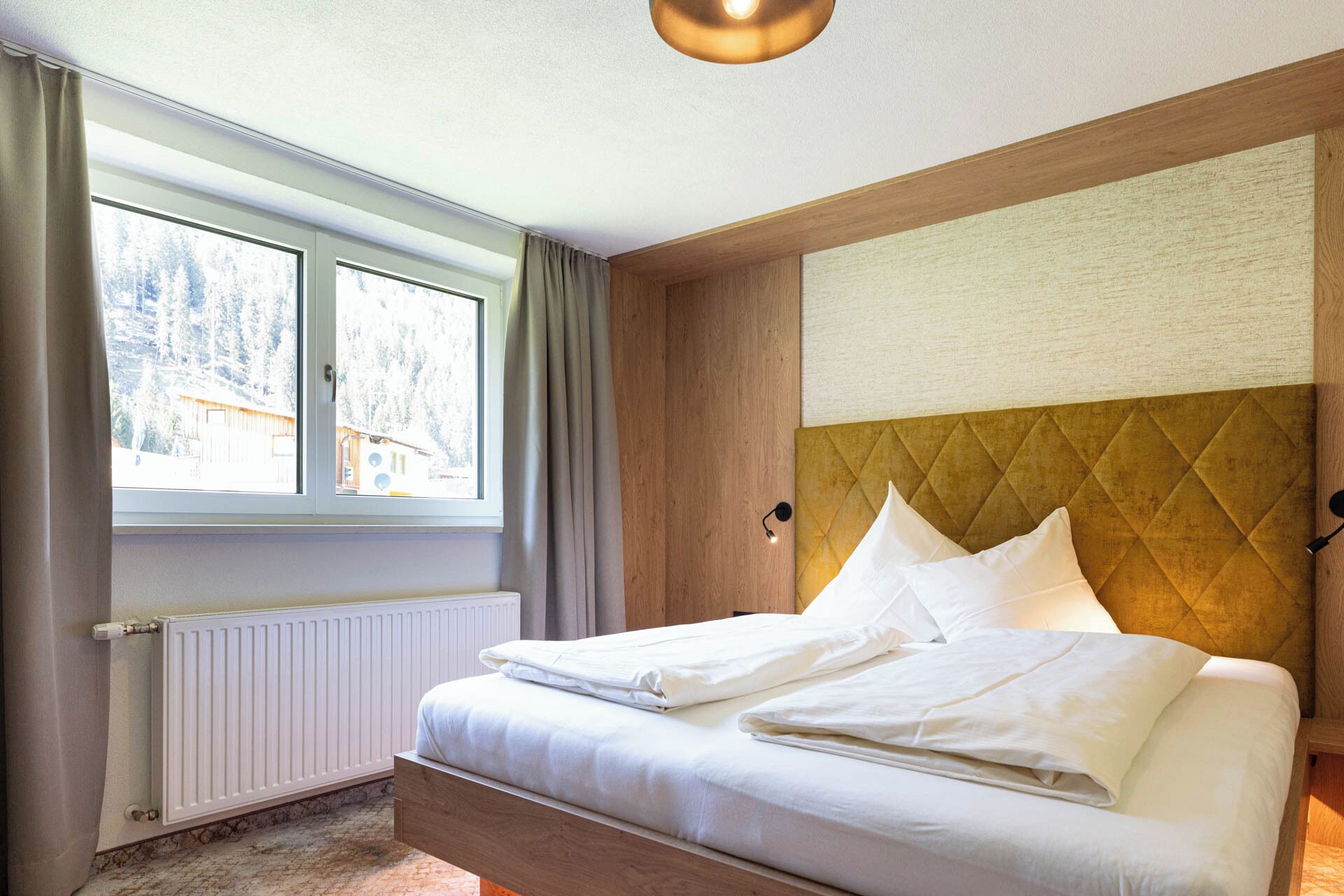 A hotel room with a bed, a window with curtains, a radiator, and a wall-mounted light fixture.