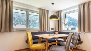 A dining area with a wooden table and chairs, a black pendant light, and a view of a snowy mountain through large windows. - Maurig Appartement (Elbigenalp)