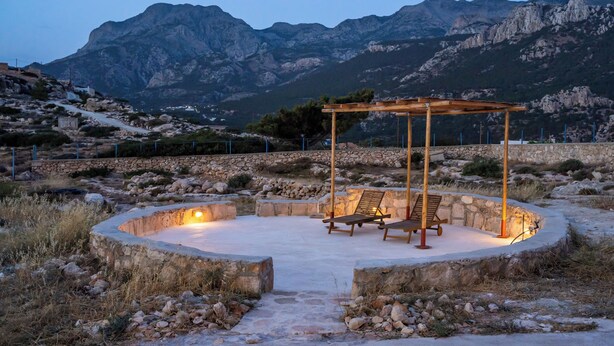 Un espacio de estar circular al aire libre con pérgola y dos sillones de descanso, rodeado de muros de piedra y un paisaje montañoso.