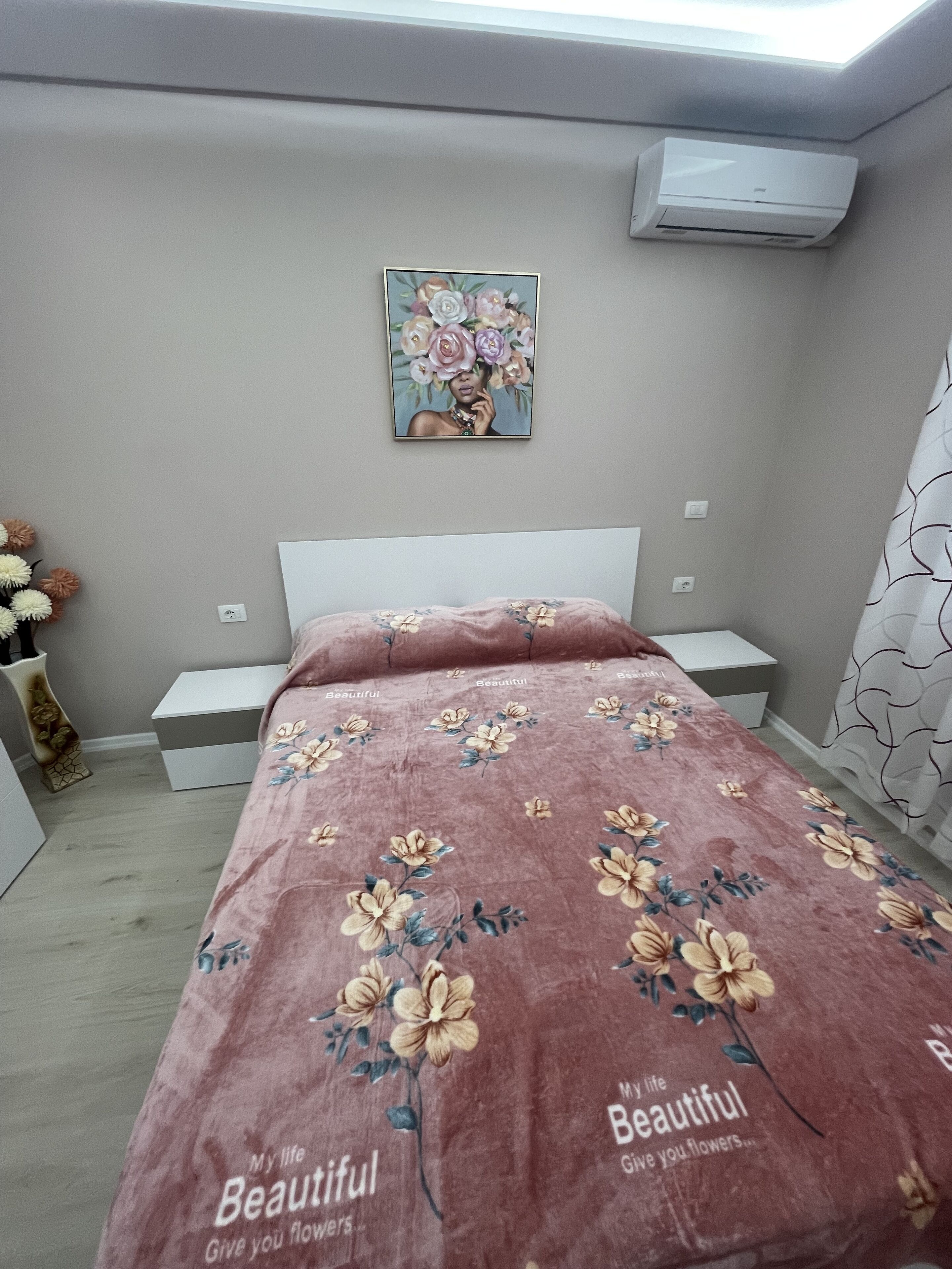 Una camera da letto con una coperta a fiori, un comodino, un tavolino notturno, un condizionatore d'aria a parete e un quadro floreale incorniciato sopra il letto.