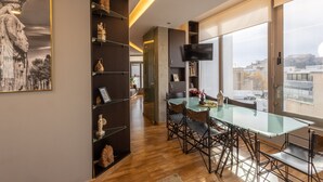 A modern dining area with a glass table and chairs, a bookshelf with decorative items, and a large window with a city view. - Athens Horizon Acropolis Views Apartment (Athens)