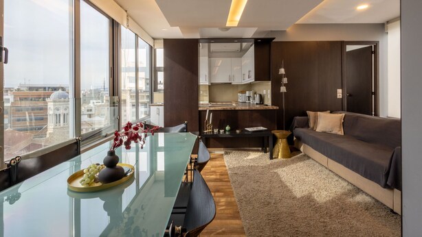 A modern living room with a glass dining table, a sofa, and a view of the city through large windows. - Athens Horizon Acropolis Views Apartment (Athens)