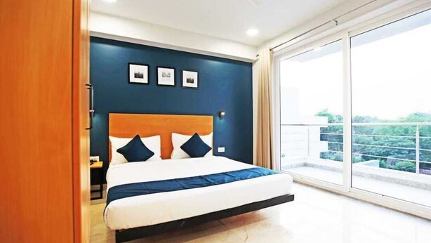 A bedroom with a bed, a nightstand, a wall-mounted shelf, and a large window with a view. - Moody moon blue (Gurugram)