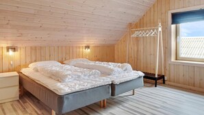 A bedroom with a sloped ceiling, wooden walls, a bed with a grey mattress, a bedside table, and a window with a view of the sky. - 5 Star Holiday Home in Lokken (Lokken)